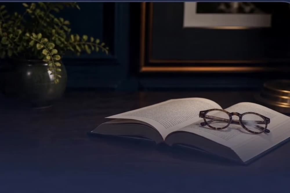 An open book and reading glasses on a dark desk beside a small green plant